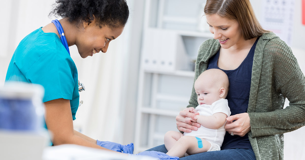 Female nurse with brown skin and turquoise scrubs smiles at a Caucasian baby