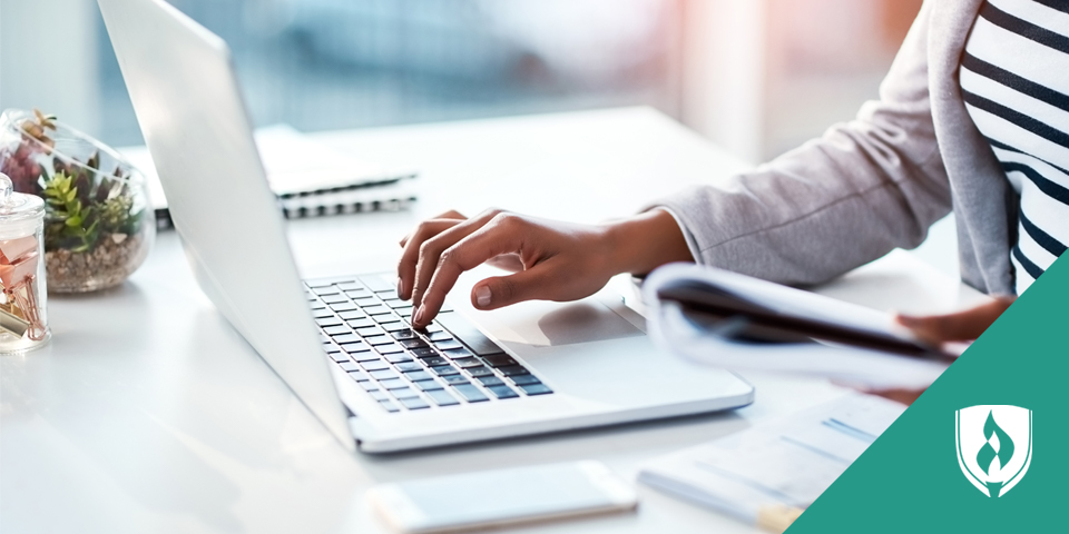 Woman typing on laptop with a notebook