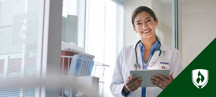 Photo of smiling nurse with white coat on and clipboard in hand.