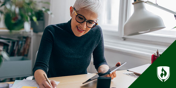 A white haired woman in a black sweater sits at her desk looking at her tablet and writing something