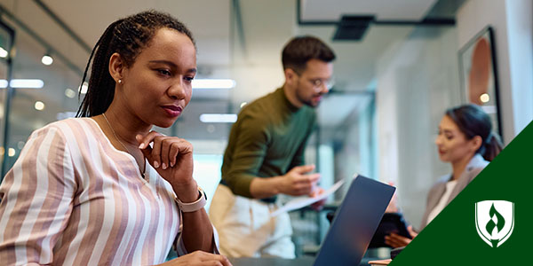 A woman in braids and a striped shirt sits at a table looking into her computer with two colleagues talking in the background