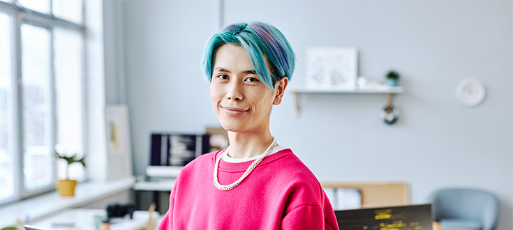 A business major with blue hair and a computer stands in their home office