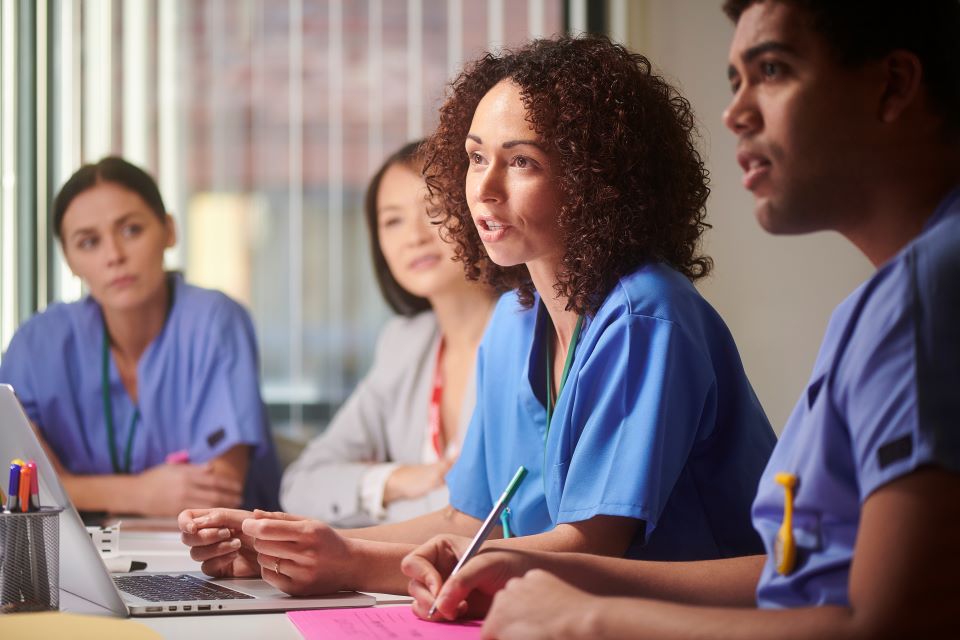 Four nursing students sit at a table, listening to a female nurse with curly brown hair