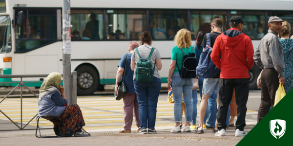 A group of people wait at a city bus stop, next to a woman sitting on a bench, her back turned to the camera