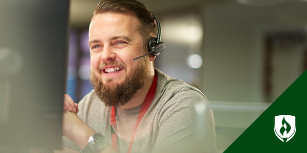 A bearded man sits smiling in front of a computer at the help desk