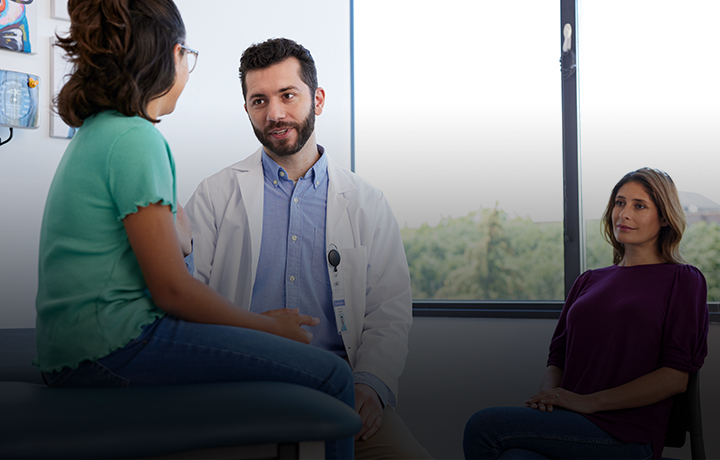 Male nurse practitioner talking with child on exam table with woman sitting in background